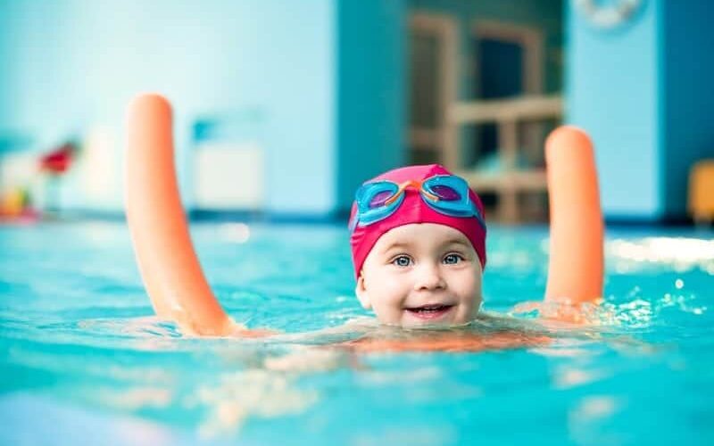 Happy little girl swimming with a noodle in indoor pool