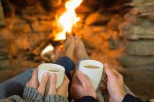 A couple warming their feet in front of the fireplace with mugs of coffee in their hands.